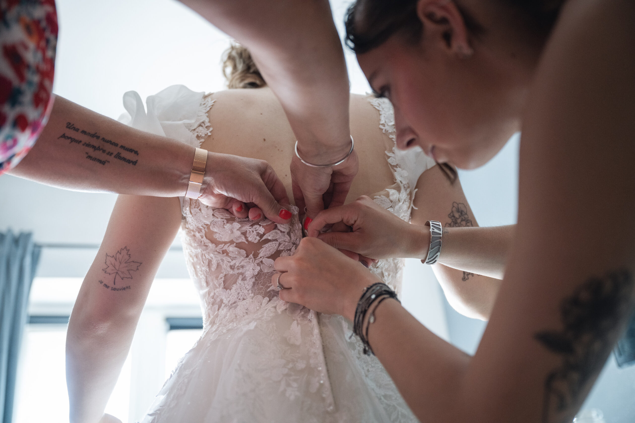Préparatifs de la mariée à Nancy, fermeture de la robe de mariée en dentelle aidée par ses proches